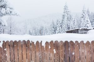 A wood fence with a snow-covered building and trees in the background.