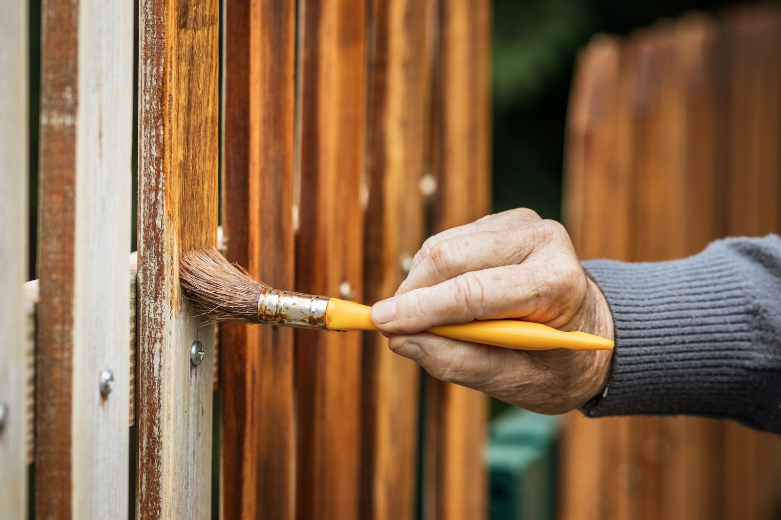 The hand of an older gentleman using a paint brush to stain a wood fence